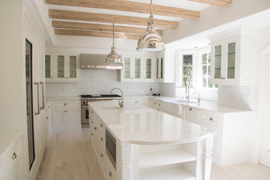Transitional u-shaped light wood floor open concept kitchen photo in Santa Barbara with a farmhouse sink, glass-front cabinets, white cabinets, white backsplash, subway tile backsplash and an island