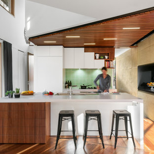 Expansive contemporary l-shaped kitchen in Melbourne with an undermount sink, flat-panel cabinets, white cabinets, green splashback, panelled appliances, medium hardwood floors, with island, brown floor, white benchtop and wood.