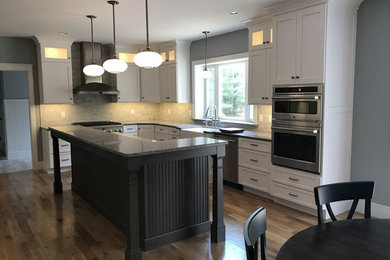 Photo of a traditional l-shaped kitchen in Boston with a belfast sink, shaker cabinets, white cabinets, granite worktops, grey splashback, stone tiled splashback, stainless steel appliances, light hardwood flooring and an island.