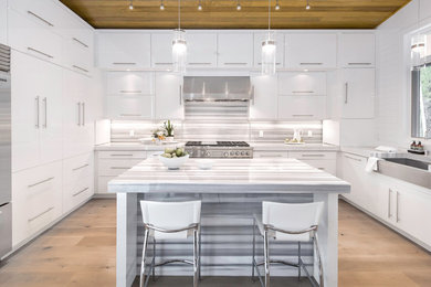 Example of a mid-sized minimalist u-shaped medium tone wood floor eat-in kitchen design in Denver with a farmhouse sink, flat-panel cabinets, white cabinets, marble countertops, gray backsplash, stone slab backsplash, stainless steel appliances and an island