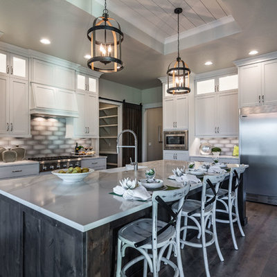 Farmhouse l-shaped dark wood floor and brown floor kitchen photo in Boise with a farmhouse sink, shaker cabinets, white cabinets, gray backsplash, stainless steel appliances, an island and white countertops