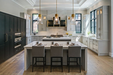 Example of a transitional u-shaped medium tone wood floor, brown floor and tray ceiling kitchen design in Chicago with an undermount sink, shaker cabinets, gray cabinets, white backsplash, stone slab backsplash, stainless steel appliances, two islands and white countertops