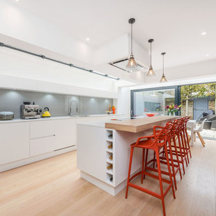 This is an example of a contemporary galley kitchen/diner in Other with flat-panel cabinets, white cabinets, wood worktops, grey splashback, black appliances, light hardwood flooring, an island and beige floors.