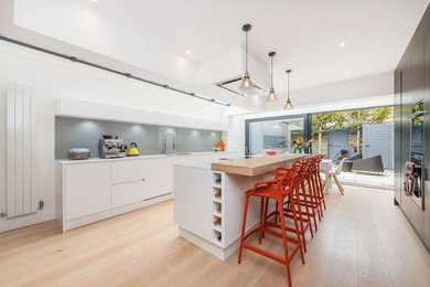 This is an example of a contemporary galley kitchen/diner in Other with flat-panel cabinets, white cabinets, wood worktops, grey splashback, black appliances, light hardwood flooring, an island and beige floors.