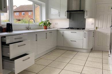 Inspiration for a medium sized contemporary l-shaped kitchen/diner in Cambridgeshire with a submerged sink, flat-panel cabinets, white cabinets, composite countertops, grey splashback, glass sheet splashback, black appliances, ceramic flooring, no island, white floors and white worktops.