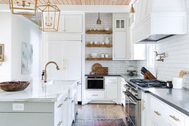 Example of a cottage l-shaped medium tone wood floor and brown floor kitchen design in Houston with a farmhouse sink, shaker cabinets, white cabinets, white backsplash, subway tile backsplash, an island and gray countertops