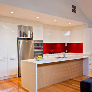 Contemporary l-shaped kitchen in Melbourne with a double-bowl sink, flat-panel cabinets, white cabinets, red splashback, glass sheet splashback, stainless steel appliances, medium hardwood floors, with island, brown floor and white benchtop.