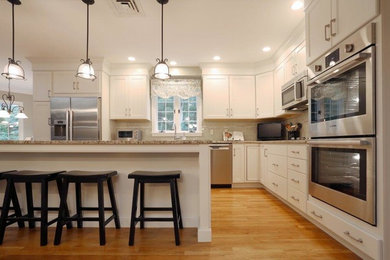 Mid-sized transitional l-shaped light wood floor open concept kitchen photo in Boston with white cabinets, granite countertops, gray backsplash, glass tile backsplash, stainless steel appliances, an island, an undermount sink and shaker cabinets