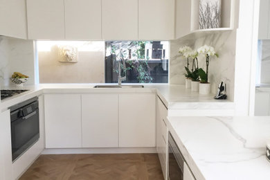 Medium sized traditional u-shaped kitchen/diner in Melbourne with a single-bowl sink, flat-panel cabinets, white cabinets, marble worktops, white splashback, marble splashback, black appliances, light hardwood flooring, an island, brown floors and white worktops.