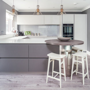 Design ideas for a contemporary u-shaped kitchen in Berkshire with a single-bowl sink, flat-panel cabinets, white cabinets, grey splashback, glass sheet splashback, black appliances, light hardwood flooring, a breakfast bar and white worktops.