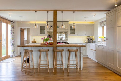 Country Küche in L-Form mit Landhausspüle, Schrankfronten mit vertiefter Füllung, beigen Schränken, Küchenrückwand in Weiß, braunem Holzboden, Kücheninsel, braunem Boden und weißer Arbeitsplatte in Hampshire