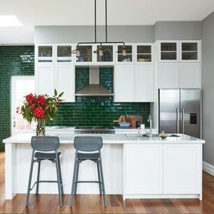 Inspiration for a transitional galley open plan kitchen in Sydney with a double-bowl sink, shaker cabinets, white cabinets, green splashback, subway tile splashback, stainless steel appliances, medium hardwood floors, with island, brown floor and white benchtop.