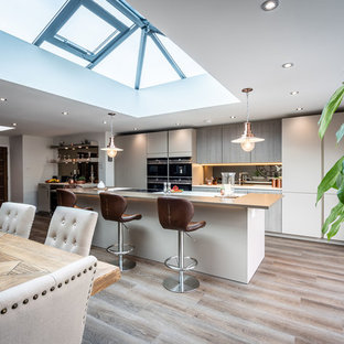 Photo of a contemporary galley kitchen/diner in Hampshire with flat-panel cabinets, beige cabinets, mirror splashback, black appliances, light hardwood flooring, an island, beige floors and white worktops.