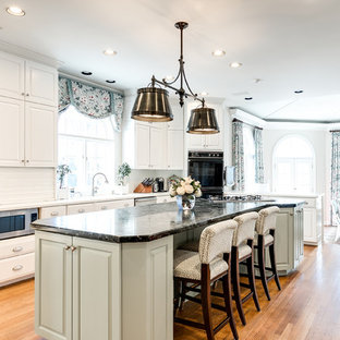 Modelo de cocina comedor tradicional con armarios con paneles con relieve, puertas de armario blancas, salpicadero blanco, electrodomésticos de acero inoxidable, suelo de madera en tonos medios, una isla y encimeras blancas