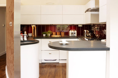Foto på ett mellanstort funkis kök, med släta luckor, vita skåp, rött stänkskydd, mellanmörkt trägolv, en halv köksö, en nedsänkt diskho, träbänkskiva och integrerade vitvaror