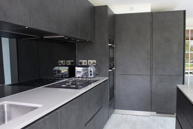 This is an example of a large contemporary kitchen in Essex with black cabinets, quartz worktops, black splashback, glass sheet splashback, black appliances, light hardwood flooring and an island.