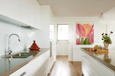 Foto på ett mellanstort funkis linjärt kök och matrum, med en dubbel diskho, släta luckor, vita skåp, bänkskiva i kvarts, vitt stänkskydd, stänkskydd i porslinskakel, rostfria vitvaror, mellanmörkt trägolv och en köksö