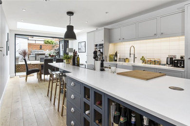 This is an example of a medium sized modern galley open plan kitchen in Surrey with a belfast sink, flat-panel cabinets, blue cabinets, composite countertops, white splashback, metro tiled splashback, black appliances, light hardwood flooring, an island, grey floors and white worktops.
