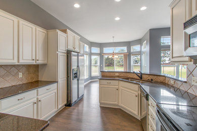 This is an example of a large traditional kitchen/diner in Milwaukee with a submerged sink, raised-panel cabinets, white cabinets, granite worktops, beige splashback, stone tiled splashback, stainless steel appliances, medium hardwood flooring, a breakfast bar, grey floors and black worktops.