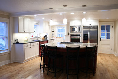 Example of a mid-sized farmhouse l-shaped light wood floor eat-in kitchen design in Portland with a drop-in sink, recessed-panel cabinets, white cabinets, quartz countertops, white backsplash, subway tile backsplash, stainless steel appliances and an island