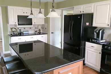 Example of a mid-sized classic u-shaped medium tone wood floor and brown floor eat-in kitchen design in Cleveland with raised-panel cabinets, white cabinets, granite countertops, white backsplash, ceramic backsplash, black appliances and an island