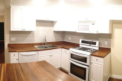 Example of a small beach style l-shaped medium tone wood floor and brown floor kitchen design in Santa Barbara with a drop-in sink, recessed-panel cabinets, white cabinets, wood countertops, white backsplash, subway tile backsplash, white appliances and an island