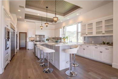 Example of a mid-sized trendy u-shaped medium tone wood floor eat-in kitchen design in Los Angeles with a double-bowl sink, raised-panel cabinets, white cabinets, granite countertops, white backsplash, mosaic tile backsplash, stainless steel appliances and two islands