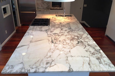 Cette image montre une cuisine américaine parallèle minimaliste avec plan de travail en marbre, une crédence blanche, un électroménager en acier inoxydable, un sol en bois brun et îlot.