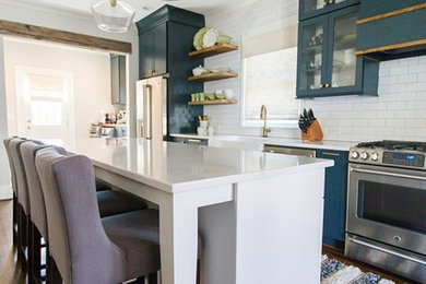 This is an example of a retro galley kitchen in Birmingham with a belfast sink, flat-panel cabinets, blue cabinets, engineered stone countertops, white splashback, metro tiled splashback, stainless steel appliances, medium hardwood flooring, an island, brown floors and white worktops.