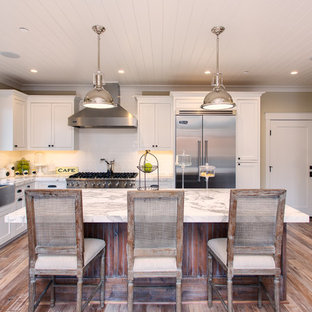 Foto de cocina en L, de estilo de casa de campo, grande, cerrada, con fregadero sobremueble, armarios estilo shaker, puertas de armario blancas, encimera de mármol, salpicadero blanco, electrodomésticos de acero inoxidable, suelo de madera en tonos medios, una isla y salpicadero de azulejos tipo metro