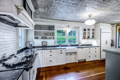 Example of a mid-sized country l-shaped medium tone wood floor enclosed kitchen design in Portland Maine with a farmhouse sink, white cabinets, soapstone countertops, white backsplash, ceramic backsplash, paneled appliances, shaker cabinets and an island