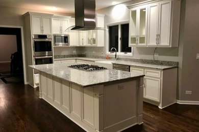 Large traditional l-shaped kitchen/diner in Chicago with a belfast sink, white cabinets, grey splashback, stainless steel appliances, dark hardwood flooring, an island, brown floors, recessed-panel cabinets and granite worktops.