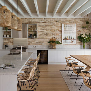 Inspiration for a beach style l-shaped eat-in kitchen in Sydney with a drop-in sink, flat-panel cabinets, white cabinets, brick splashback, stainless steel appliances, light hardwood floors and with island.