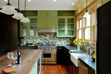 Example of a classic l-shaped kitchen design in Seattle with a farmhouse sink, glass-front cabinets, green cabinets, green backsplash, subway tile backsplash, stainless steel appliances and wood countertops