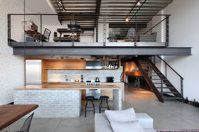 Photo of a small urban galley open plan kitchen in Seattle with concrete flooring, an island, flat-panel cabinets, light wood cabinets, wood worktops, grey splashback, brick splashback, stainless steel appliances, a submerged sink and grey floors.