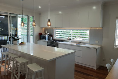 Large contemporary galley open plan kitchen in Brisbane with a submerged sink, flat-panel cabinets, white cabinets, engineered stone countertops, grey splashback, glass sheet splashback, stainless steel appliances, medium hardwood flooring, multiple islands, brown floors and white worktops.