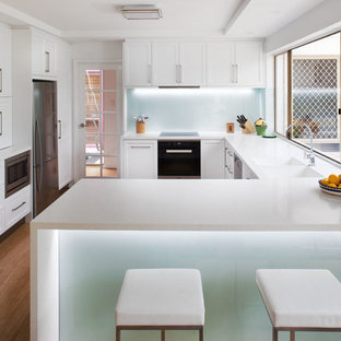 This is an example of a mid-sized transitional u-shaped kitchen in Perth with an integrated sink, recessed-panel cabinets, white cabinets, solid surface benchtops, white splashback, glass sheet splashback, stainless steel appliances, bamboo floors and a peninsula.