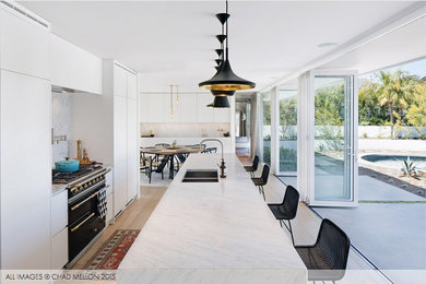 Example of a mid-sized minimalist light wood floor and beige floor open concept kitchen design in Orange County with a farmhouse sink, flat-panel cabinets, white backsplash, marble backsplash, stainless steel appliances and an island