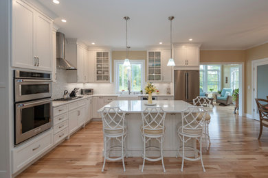 Inspiration for a large transitional l-shaped medium tone wood floor and brown floor eat-in kitchen remodel in Boston with a farmhouse sink, shaker cabinets, white cabinets, quartz countertops, white backsplash, mosaic tile backsplash, stainless steel appliances, an island and white countertops