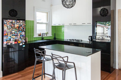 Example of a trendy l-shaped medium tone wood floor and brown floor kitchen design in Melbourne with a drop-in sink, flat-panel cabinets, black cabinets, green backsplash, an island and black countertops