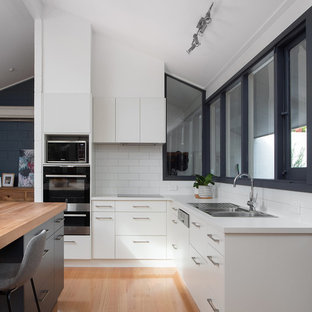 Design ideas for a contemporary l-shaped kitchen in Hobart with a drop-in sink, flat-panel cabinets, white cabinets, white splashback, stainless steel appliances, medium hardwood floors, with island, brown floor and white benchtop.
