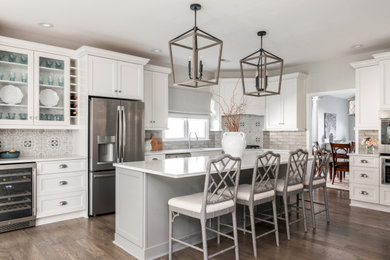 This is an example of a medium sized farmhouse l-shaped kitchen/diner in Charlotte with a submerged sink, shaker cabinets, white cabinets, engineered stone countertops, grey splashback, metro tiled splashback, stainless steel appliances, an island, white worktops, dark hardwood flooring and brown floors.