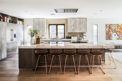 This is an example of a large contemporary l-shaped open plan kitchen in Los Angeles with flat-panel cabinets, an island, distressed cabinets, composite countertops, a submerged sink, beige splashback, stone tiled splashback, stainless steel appliances and light hardwood flooring.