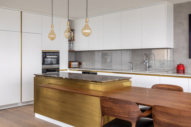 Large contemporary l-shaped kitchen in London with a double-bowl sink, flat-panel cabinets, white cabinets, grey splashback, glass sheet splashback, stainless steel appliances, medium hardwood flooring, an island, brown floors and white worktops.