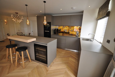This is an example of a medium sized modern l-shaped open plan kitchen in London with an integrated sink, flat-panel cabinets, grey cabinets, laminate countertops, metallic splashback, mirror splashback, black appliances, light hardwood flooring, an island and grey worktops.
