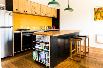 Bild på ett funkis parallellkök, med en nedsänkt diskho, släta luckor, svarta skåp, gult stänkskydd, stänkskydd i tunnelbanekakel, rostfria vitvaror, mellanmörkt trägolv och en köksö