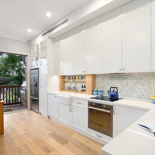 This is an example of a transitional l-shaped eat-in kitchen in Sydney with white cabinets, blue splashback, stainless steel appliances, a farmhouse sink, shaker cabinets, light hardwood floors, a peninsula, white benchtop and mosaic tile splashback.