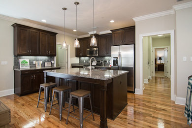 Example of a mid-sized arts and crafts l-shaped medium tone wood floor eat-in kitchen design in Atlanta with a drop-in sink, recessed-panel cabinets, dark wood cabinets, granite countertops, gray backsplash, stainless steel appliances, an island and glass tile backsplash