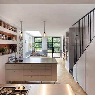 Exemple d'une cuisine parallèle tendance de taille moyenne avec un plan de travail en inox, un îlot central, un évier encastré, un placard à porte plane, des portes de placard blanches, un sol en bois clair, un sol beige et un plan de travail noir.
