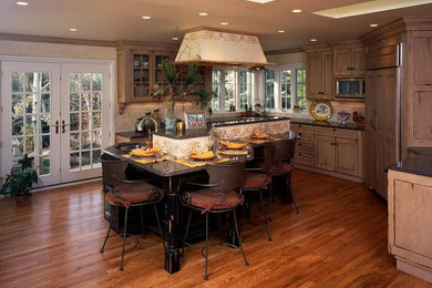 Photo of a medium sized traditional u-shaped kitchen/diner in Denver with a submerged sink, recessed-panel cabinets, distressed cabinets, granite worktops, beige splashback, travertine splashback, integrated appliances, dark hardwood flooring, an island and brown floors.